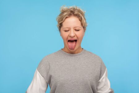 Naughty woman with short hair in sweatshirt standing with closed eyes and sticking out tongue, making derisive goofy face, being disobedient unruly. indoor studio shot isolated on blue backgroundの写真素材