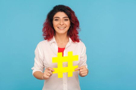 Trendy blogs. Portrait of charming woman with fancy red hair holding big hashtag symbol and smiling...