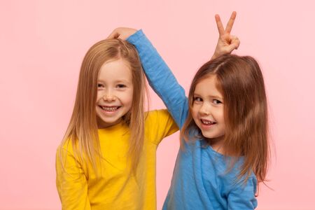 Happy carefree childhood, friendship. Portrait of two positive cute little girls showing bunny ears to each other, having fun, playing games together. indoor studio shot isolated on pink backgroundの写真素材