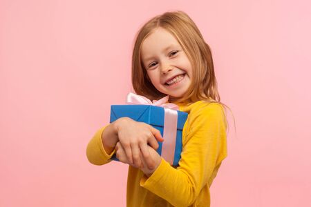 Delighted charming little girl embracing gift box and smiling joyfully to camera, satisfied with...
