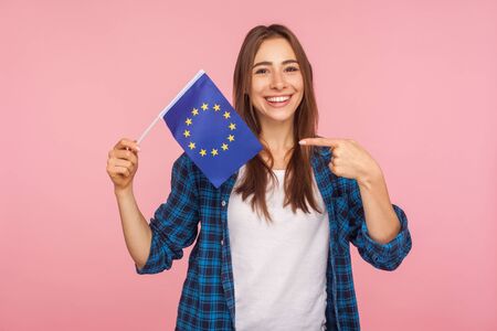 Portrait of positive friendly woman in checkered shirt smiling broadly and pointing at European Union flag, symbol of Europe, EU association, community. indoor studio shot isolated on pink backgroundの写真素材