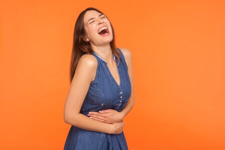 Hilarious laughter. Joyful brunette woman in denim dress holding her belly bursting into laughing loud, being hysterical at joke, funny anecdote. indoor studio shot isolated on orange backgroundの写真素材