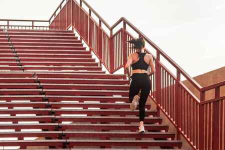 Back view of runner athlete, fit brunette girl in black sportswear, tight pants and top, running up...