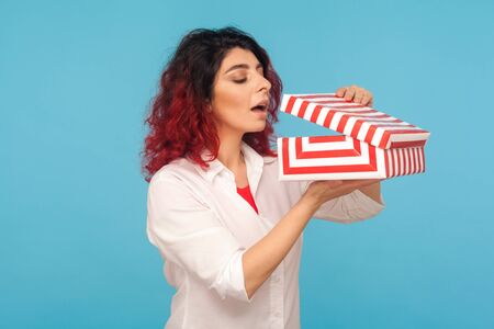 Beautiful hipster woman with fancy red hair eagerly opening gift box and peeking inside with...