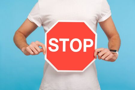 Closeup of man in white t-shirt holding Stop symbol in hands, warning with red traffic sign, concept of ban, denial, forbidden way, prohibited to go. indoor studio shot isolated on blue backgroundの写真素材
