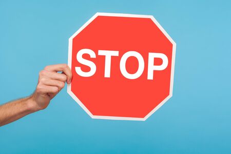 Closeup of male hand holding Stop symbol, showing red traffic sign, warning about problems, concept of ban, denial, prohibited to go, forbidden way. indoor studio shot isolated on blue backgroundの写真素材