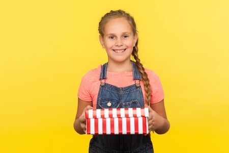 Portrait of excited glad little girl with braid in denim overalls holding gift box and smiling to...