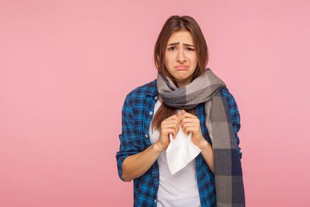 Seasonal influenza. Portrait of ill girl in checkered shirt wrapped in scarf holding napkin, suffering runny nose and sore throat, symptoms of flu, viral infection. indoor studio shot, pink backgroundの写真素材