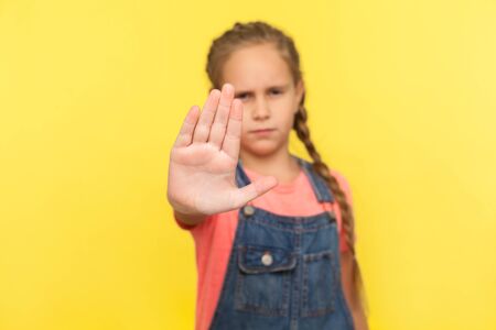 No, rejection sign. Portrait of unhappy little girl in denim overalls showing stop gesture, warning to go, looking angry dissatisfied, denial concept. indoor studio shot isolated on yellow backgroundの写真素材