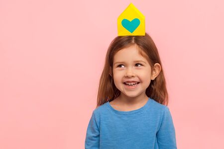Charming brunette little girl looking at paper house with heart on her head and laughing, adorable child dreaming of home, adoption and loving family. indoor studio shot isolated on pink backgroundの写真素材
