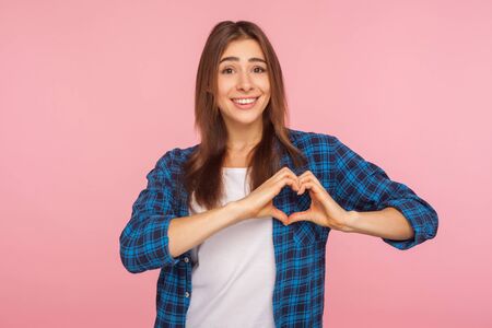 Portrait of lovely romantic girl in checkered shirt showing heart shape with hands and smiling at...