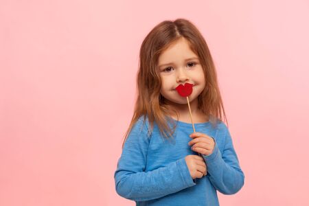 Portrait of lovely cute brunette little girl covering mouth with red paper lips or heart shape, kindly looking at camera, fashion and beauty concept. indoor studio shot isolated on pink backgroundの写真素材