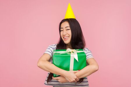 Portrait of pleased happy girl with party cone on head embracing big gift box and smiling at camera,...
