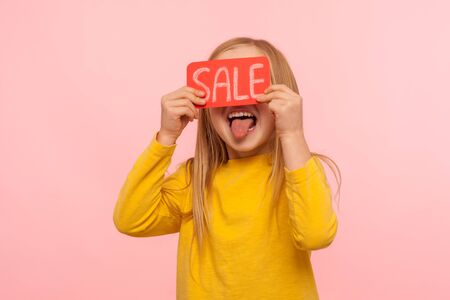 Weekend discounts! Carefree adorable little girl covering eyes with Sale word and showing tongue at camera, excited about low prices in kid store, Black Friday. indoor studio shot, pink backgroundの写真素材