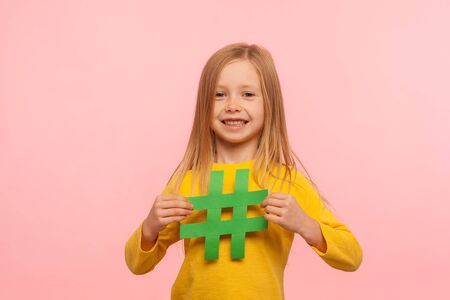Popular kids content. Portrait of joyful cute adorable little girl holding hashtag symbol and looking at camera with toothy smile, showing hash sign. indoor studio shot isolated on pink backgroundの写真素材