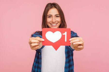 Portrait of happy blogger girl in checkered shirt smiling and showing like icon, heart button to...