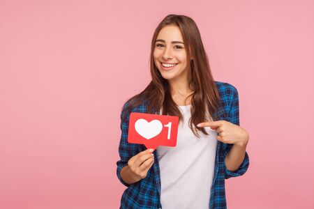 I like this content! Charming positive girl in checkered shirt smiling playfully and holding heart icon, social media button to enjoy trendy interesting blog. studio shot isolated on pink backgroundの写真素材