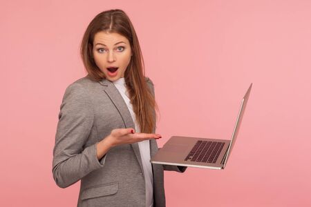 Portrait of astonished businesswoman holding laptop and looking at camera with amazed face, using computer to study, working online, shocked by application. studio shot isolated on pink backgroundの写真素材