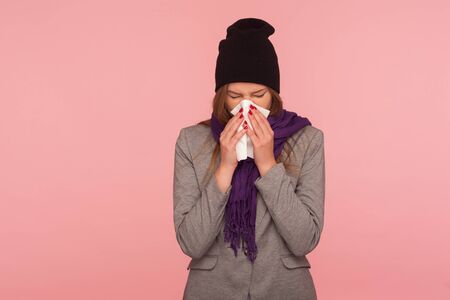 Seasonal influenza. Portrait of flu-sick woman in warm hat and scarf blowing nose in tissue,...