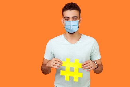 Promotion in social media. Portrait of happy brunette man with surgical medical mask smiling and holding big hashtag sign, sharing viral content. indoor studio shot isolated on orange backgroundの写真素材