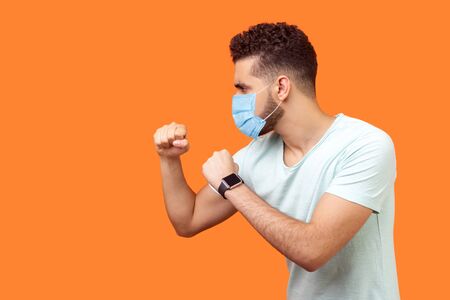 Boxing and self-defence. Side view of aggressive man with surgical medical mask keeping fists clenched, ready for fight. empty copy space on left for text. studio shot isolated on orange backgroundの写真素材