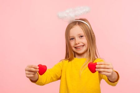 Portrait of cute angelic happy little girl with halo over head holding toy hearts and looking at...