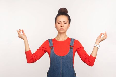 Portrait of calm stylish pretty girl with hair bun in denim overalls holding hands in mudra gesture...