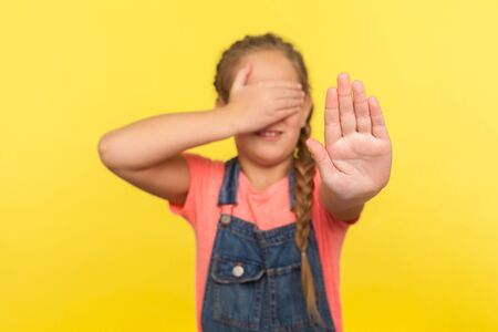 Portrait of frightened little girl with braid in denim overalls covering eyes with hand and showing stop gesture, child feeling shame or fear to watch forbidden content. indoor studio shot, isolatedの写真素材