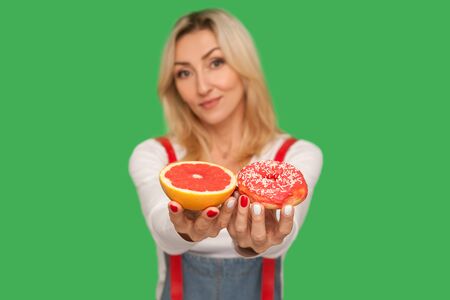 Choose healthy fruit or junk food. Portrait of positive woman showing sweet doughnut and fresh juicy grapefruit, offering choice, carbs and glucose in diet. studio shot isolated on green backgroundの写真素材