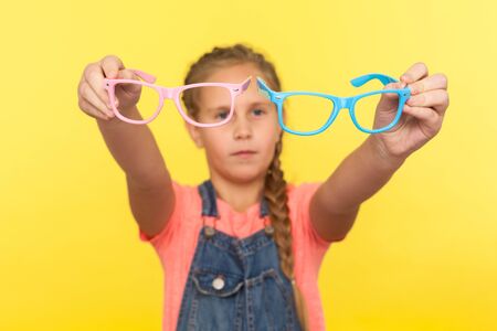 Eyesight and eye health. Portrait of little girl holding pink and blue eyeglasses and expressing doubts, child choosing spectacles in optics store. indoor studio shot isolated on yellow backgroundの写真素材