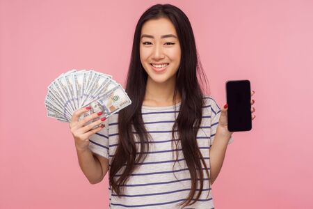 App for online mobile payment. Portrait of happy brunette girl in striped t-shirt holding money, cell phone and looking at camera with toothy smile. indoor studio shot isolated on pink backgroundの写真素材