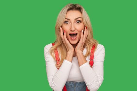 Portrait of excited amazed adult blond woman touching face and shouting in surprise, shocked by unexpected win, crazy news. indoor studio shot isolated on green backgroundの写真素材