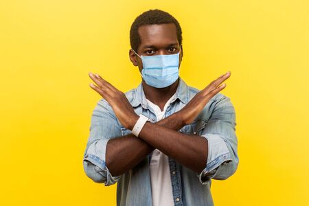 Portrait of absolutely convinced man i with surgical medical mask standing crossing hands, showing x sign meaning stop, there is no way, finish. indoor studio shot isolated on yellow backgroundの写真素材