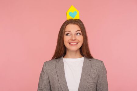 Real estate agency. Cheerful attractive woman in business suit jacket standing with paper house on her head, advertising realtor services, house purchase. studio shot isolated on pink backgroundの写真素材