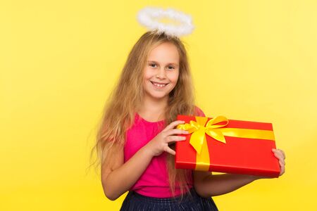 Portrait of adorable glad child girl with long blond hair and angelic halo holding gift box and smiling to camera, little angel greeting with holidays. indoor studio shot isolated on yellow backgroundの写真素材
