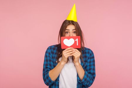 Internet trends. Portrait of positive girl with party cone on head holding social media heart like...