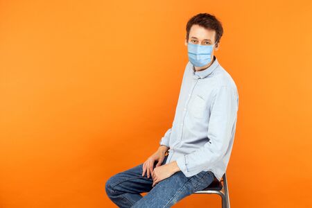 Portrait of young worker man with surgical medical mask sitting and looking at camera with serious face. health care and medicine concept. indoor studio shot isolated on orange background.の写真素材