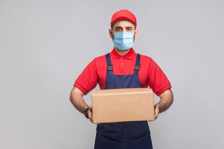 Delivery on quarantine. Portrait of young man with surgical medical mask in blue uniform and red t-shirt standing and holding the cardboard box on grey background. Indoor, studio shot, isolated,の写真素材