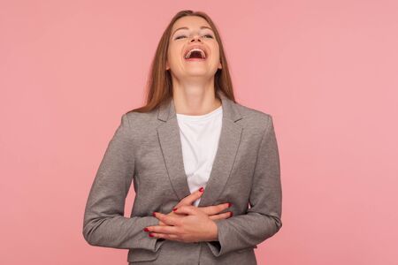 Portrait of happy carefree young woman in business suit holding her belly and laughing loud at joke, amused by funny anecdote, enjoying positive emotions. studio shot isolated on pink backgroundの写真素材