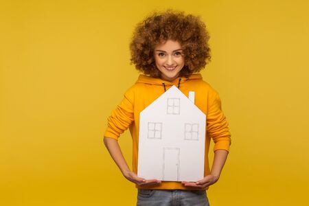 Insurance, property purchase. Joyful bright pretty woman with fluffy curly hair in hoody holding paper house and looking at camera with toothy smile. indoor studio shot isolated on yellow backgroundの写真素材
