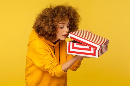 Portrait of funny curious nosy curly-haired woman in urban style hoodie looking into gift box, unwrapping present and peeking inside with interest. indoor studio shot isolated on yellow backgroundの写真素材