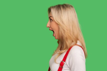Wow, unbelievable! Side view of astonished adult blond woman standing with wide open mouth and shouting in surprise, shocked expression. indoor studio shot isolated on green background, copy spaceの写真素材
