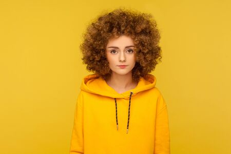 Portrait of calm serious curly-haired young beautiful woman in urban style hoodie standing, looking at camera with focused concentrated expression. indoor studio shot isolated on yellow backgroundの写真素材