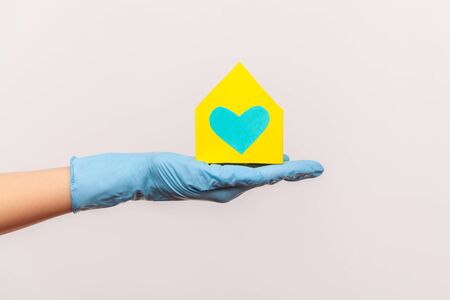 Profile side view closeup of human hand in blue surgical gloves holding yellow paper house exterior in hand with love and care. indoor, studio shot, isolated on gray background.の写真素材