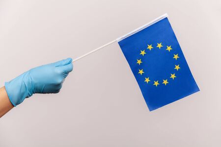 Profile side view closeup of human hand in blue surgical gloves holding Eroupean union flag. indoor,...