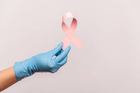 Profile side view closeup of human hand in blue surgical gloves shwoing and holding pink ribbon, symbol of breast cancer. indoor, studio shot, isolated on gray background.の写真素材