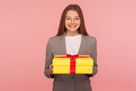 Portrait of joyful happy elegant businesswoman in suit jacket holding present and smiling, celebrating anniversary, got bonus gift for her success at work, promotion. studio shot, pink backgroundの写真素材