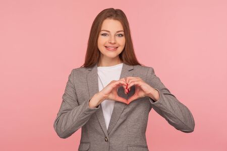 Portrait of charming friendly young woman in business suit smiling to camera and showing love heart gesture, symbol of generosity, care and hope. indoor studio shot isolated on pink backgroundの写真素材