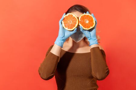 Woman in protective gloves and face mask covering eyes with orange fresh fruits, natural vitamins for immune system and flu treatment, healthy nutrition during quarantine. studio shot, isolatedの写真素材