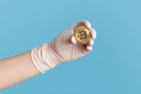 Profile side view closeup of human hand in white surgical gloves holding and showing bitcoin symbol in hand. indoor, studio shot, isolated on blue background.の写真素材